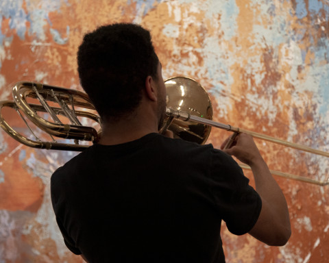 A student plays a trombone towards Jose Parla's mural which can be seen in the background as an array of orange and blue hues 